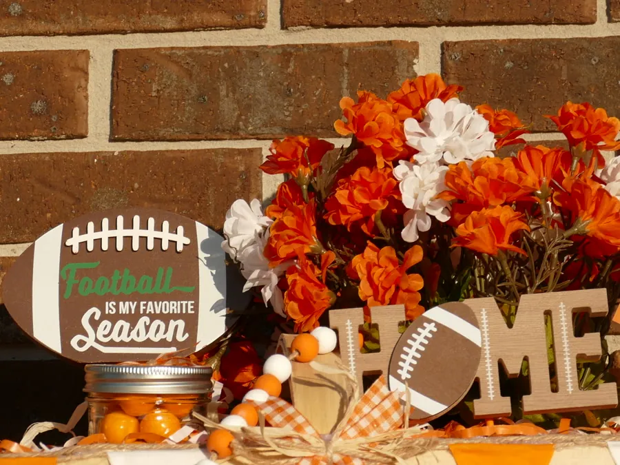 Game-day basket with orange and white florals, a wooden HOME sign with a football, an orange gingham bow, and a pennant banner across the basket weave.