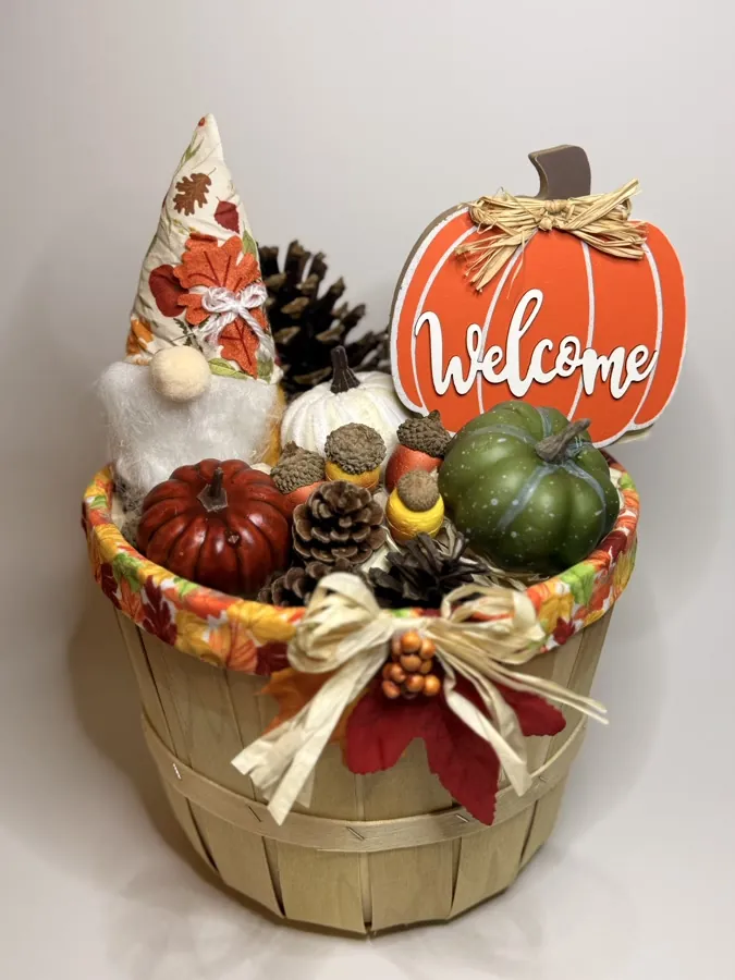 Wooden bushel basket rimmed in autumn leaf fabric, holding a fall gnome, rust and green pumpkins, painted acorns, and pinecones, with an orange wooden welcome sign standing at the back.
